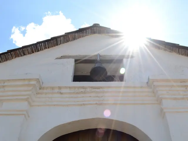 In this image we can see a parish building which has wooden door, bell and top of the image there...